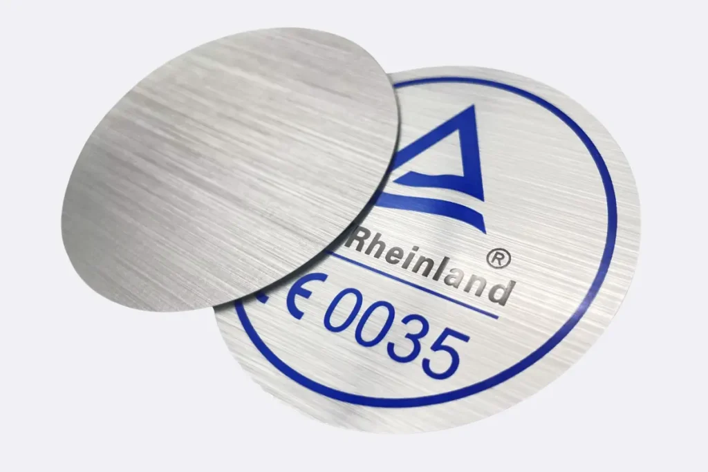 Close-up of a round brushed metallic silver sticker with a distinct grain texture, showing a CE Rheinland certification logo.